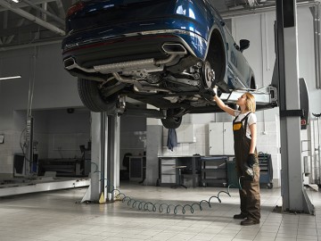 Professional female mechanic standing near automobile lifted on bridge, observing and checking wheel. Young woman looking up at brake discs. Girl wearing in coveralls.