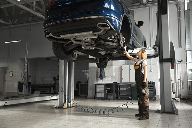 Professional female mechanic standing near automobile lifted on bridge, observing and checking wheel. Young woman looking up at brake discs. Girl wearing in coveralls.